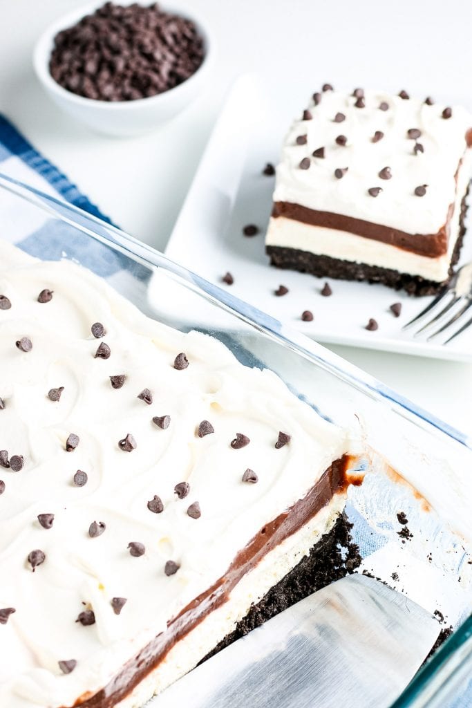 Glass pan with chocolate lasagna with a row gone. Plate with one piece on it in background.