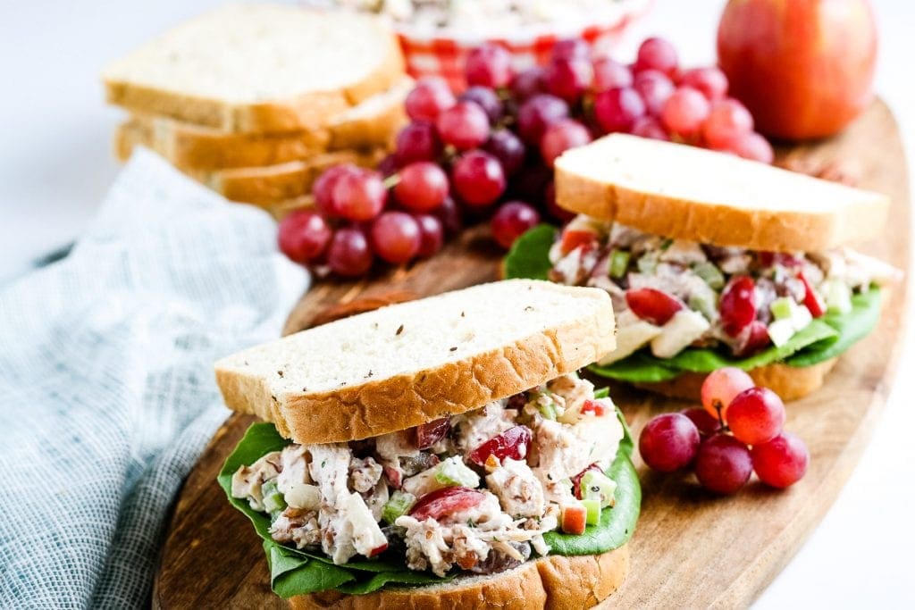 Two Chicken Salad Sandwiches on a wood cutting board with grapes and bread in background.