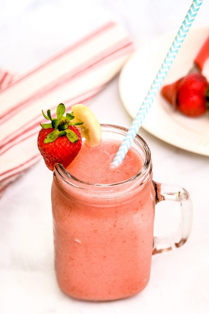 Mason Jar Mug with Strawberry Bananas Smoothie in it. Garnished with a blue and white striped paper straw, strawberry and banana slice on rim.