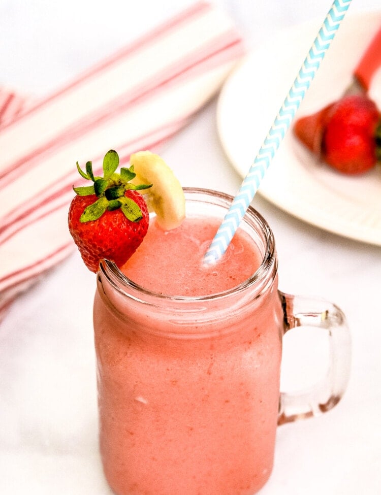 Mason Jar Mug with Strawberry Bananas Smoothie in it. Garnished with a blue and white striped paper straw, strawberry and banana slice on rim.