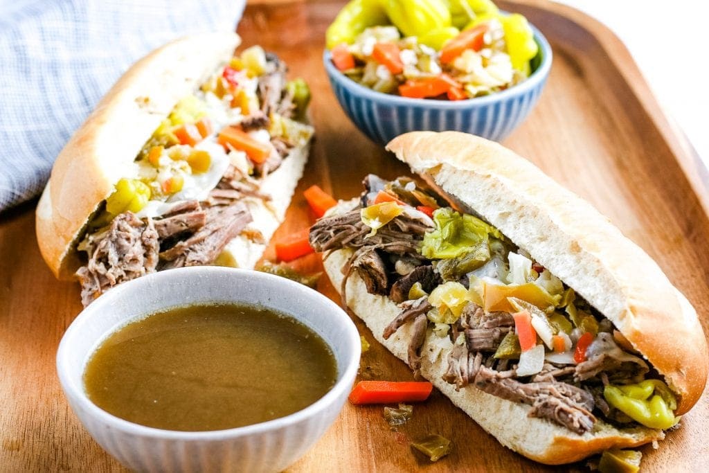 Wood cutting board with Italian Beef Sandwiches, a white bowl of beef juices from slow cooker and peppers in blue bowl in background
