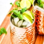 Copper mug full of ice and moscow mule garnished with lime wedges and mint on wooden cutting board.