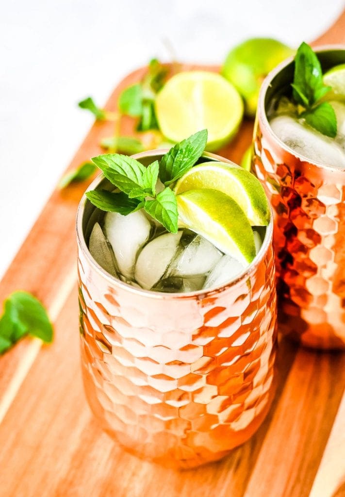 Copper mug full of ice and moscow mule garnished with lime wedges and mint on wooden cutting board.