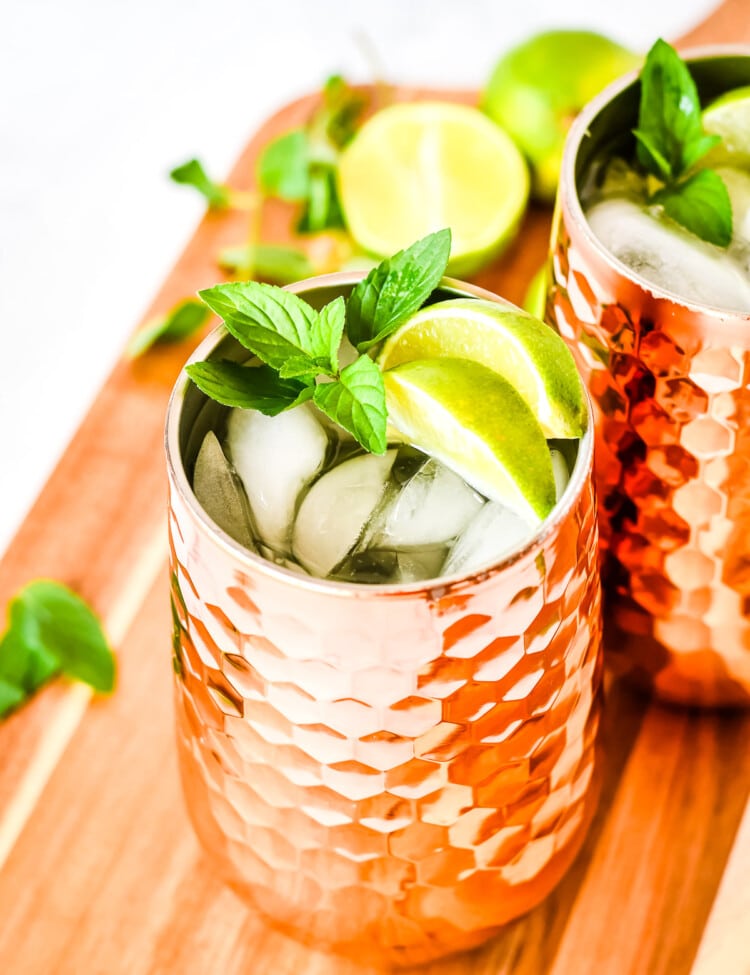 Copper mug full of ice and moscow mule garnished with lime wedges and mint on wooden cutting board.