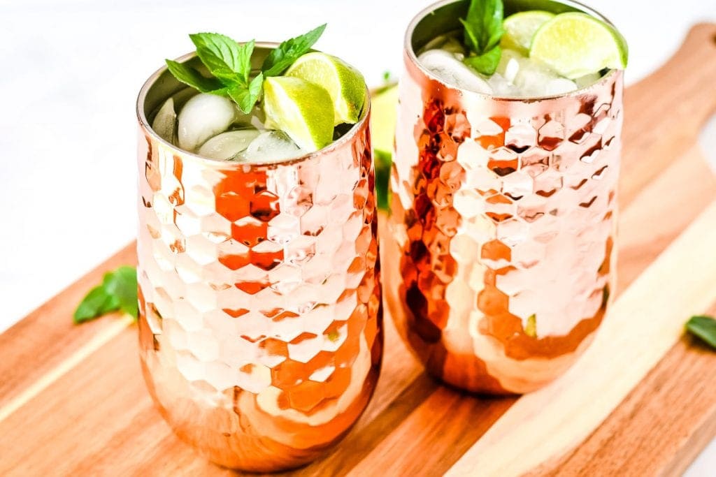 Two copper mugs with moscule mule in them garnished with lime wedges and mint on wooden board.