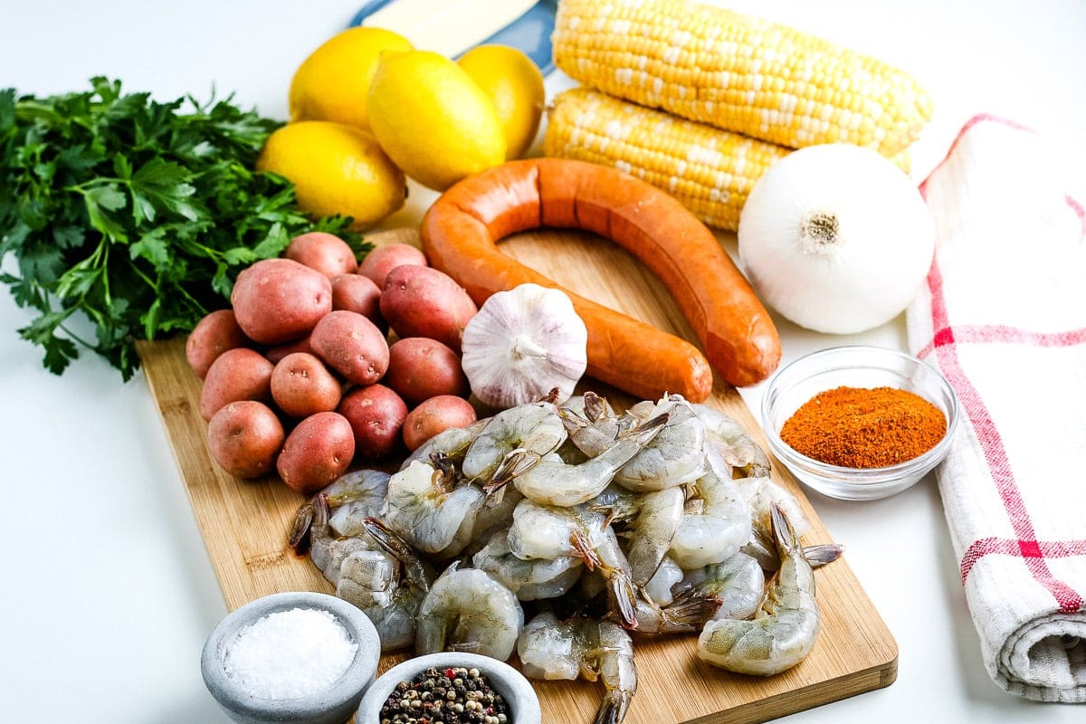 Wood cutting board showing ingredients for shrimp boil like raw shrimp, baby red potatoes, corn on the cob, smoked sausage and seaonings.