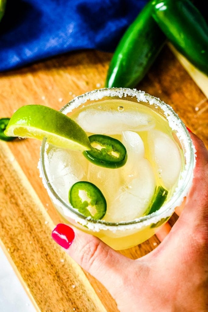 Hand holding a glass of spicy margarita that is rimmed with salt, sliced jalapenos and a lime wedge garnish