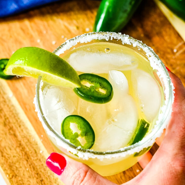 Hand holding a glass of spicy margarita that is rimmed with salt, sliced jalapenos and a lime wedge garnish