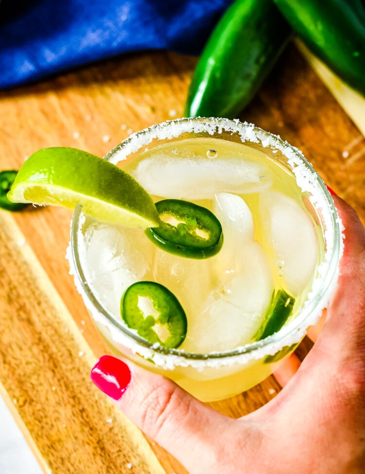 Hand holding a glass of spicy margarita that is rimmed with salt, sliced jalapenos and a lime wedge garnish