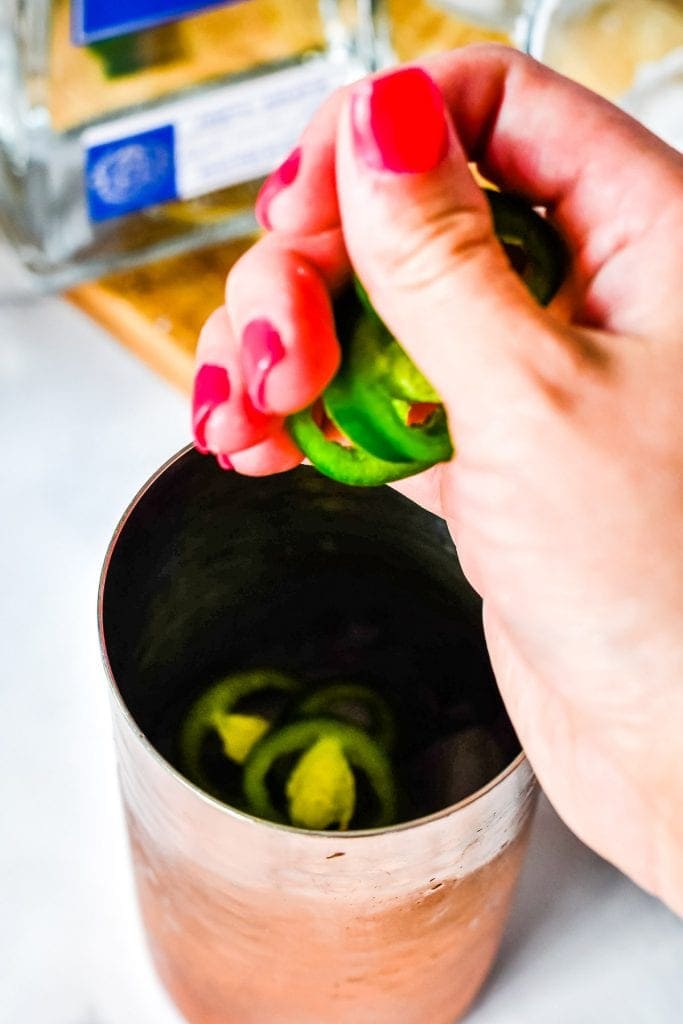 Hand putting sliced jalapenos in copper cocktail shaker