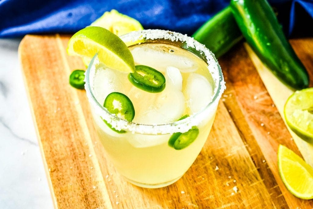 Spicy Margarita sitting on wooden cutting board in a salt rimmed glass that's garnished with a lime wedge.
