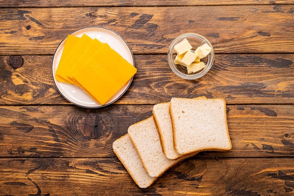 Overhead image of ingredients for recipe including cheese slices on a plate, butter in a glass dish and slices of bread.