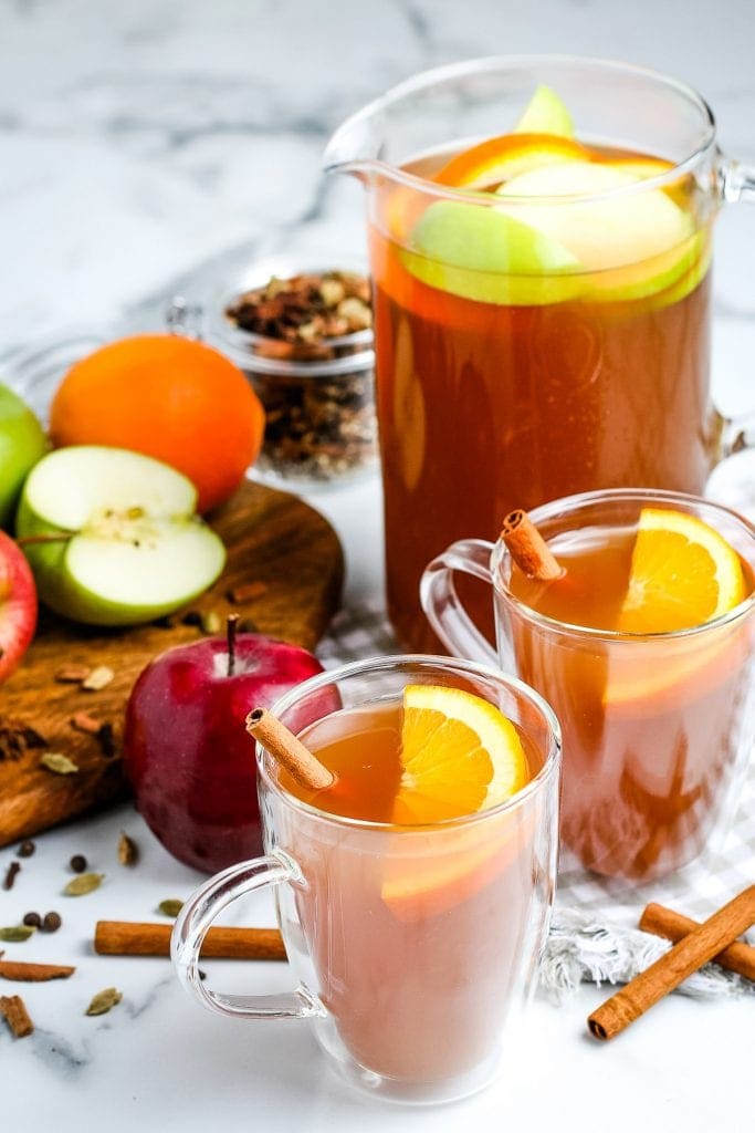 Apple Cider in glass mugs garnished with orange slices and cinnamon sticks. Pitcher in background that's full.