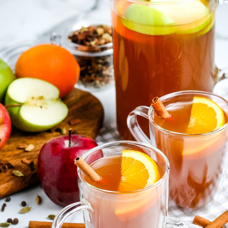 Apple Cider in glass mugs garnished with orange slices and cinnamon sticks. Pitcher in background that's full.