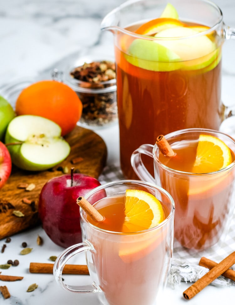 Apple Cider in glass mugs garnished with orange slices and cinnamon sticks. Pitcher in background that's full.