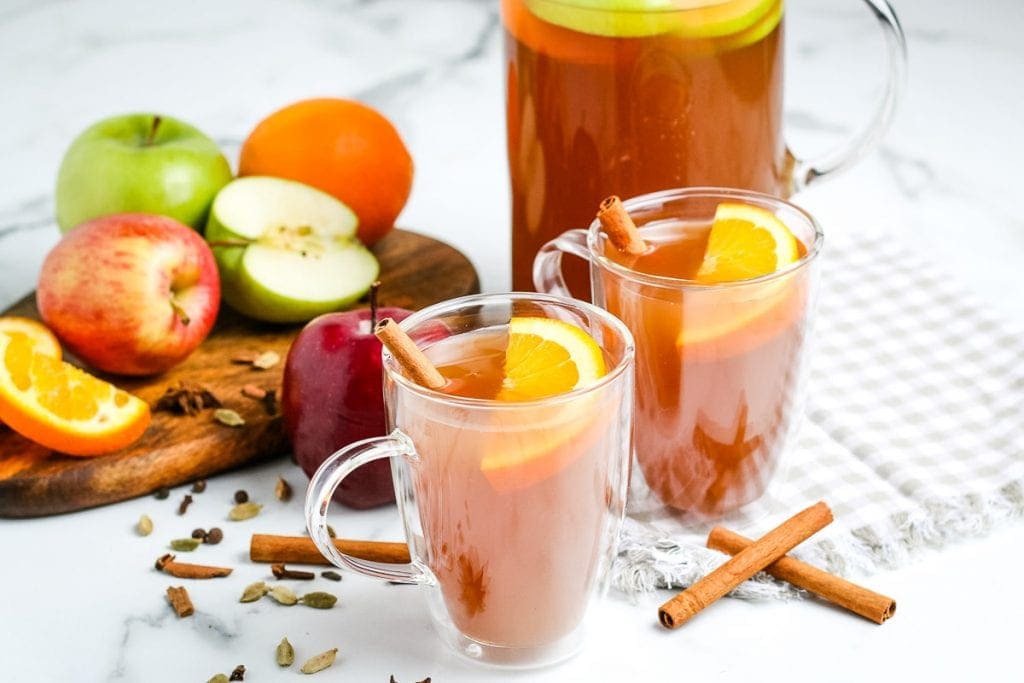 Two glass mugs with apple cider. Garnished with cinnamon sticks and oranges. Pitcher and sliced apples and oranges in background.