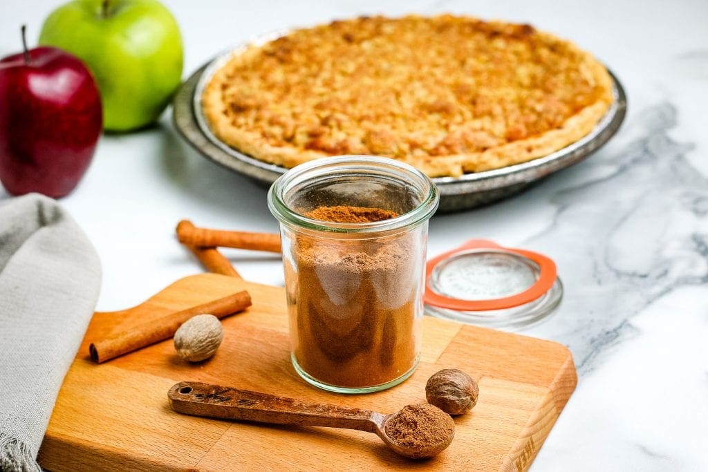 Homemade Apple Pie Spice in a glass container sitting on wooden cutting board and apple pie in background.