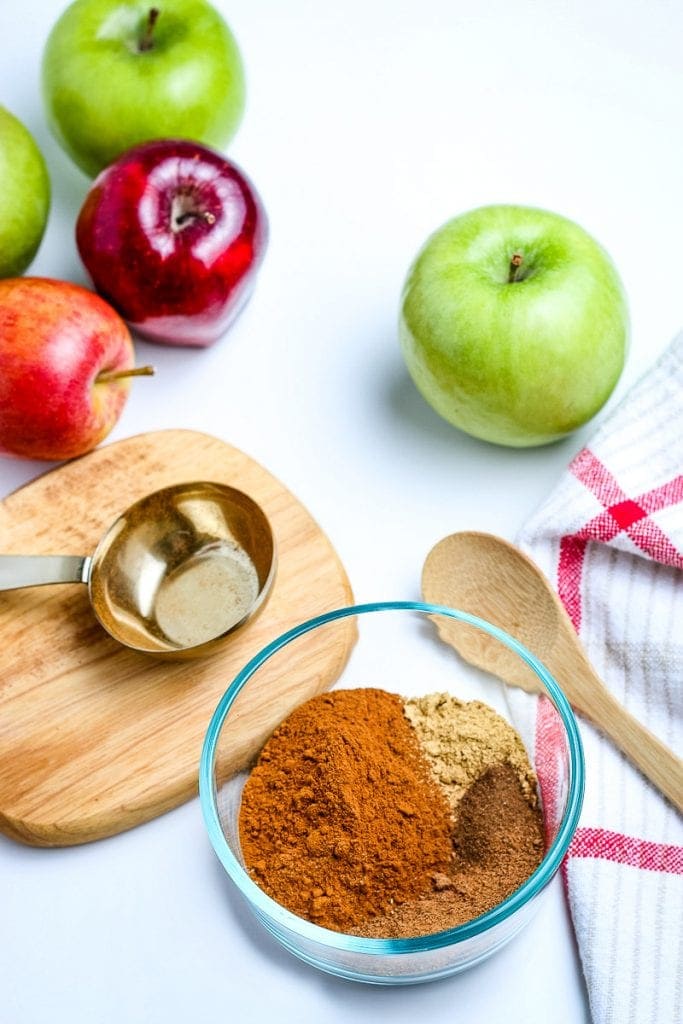 Small glass bowl with apple pie spice ingredients in it before mixing. Apples and measuring spoons in background.