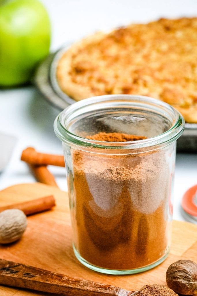 Glass jar on light wooden board with apple pie spice in it and pie in background.