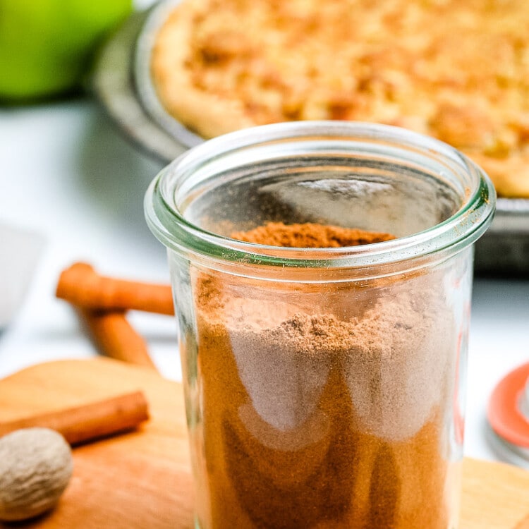Glass jar on light wooden board with apple pie spice in it and pie in background.