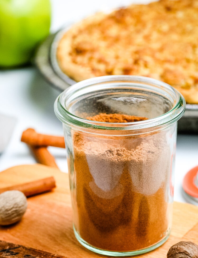 Glass jar on light wooden board with apple pie spice in it and pie in background.