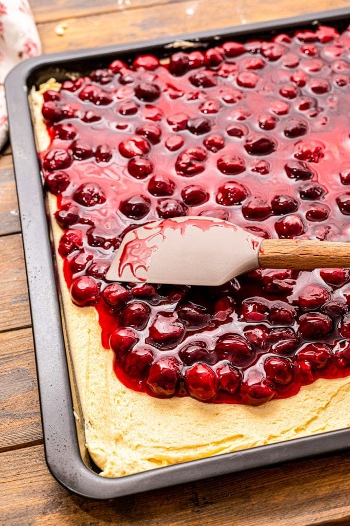 Spatula spreading cherry pie filling over crust batter in pan