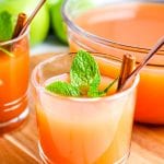 Glass of Instant Pot Apple Cider with a bowl of it in background sitting on wooden background.