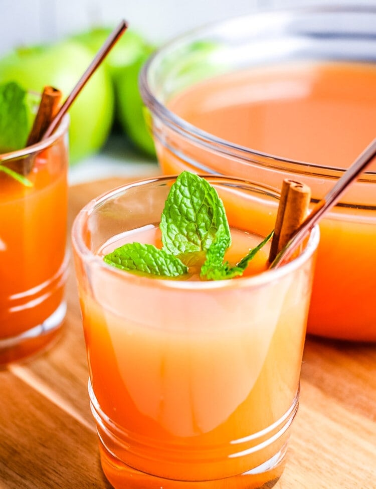 Glass of Instant Pot Apple Cider with a bowl of it in background sitting on wooden background.