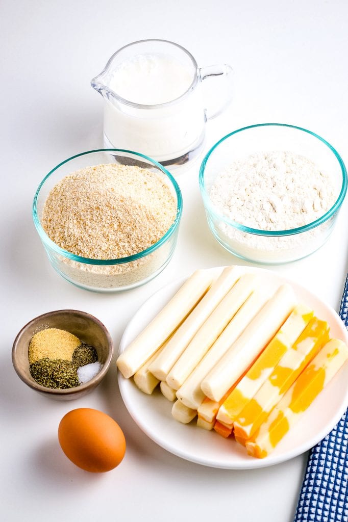 Ingredients for Mozzarella sticks including an egg, seasonings, plate of cheese sticks, bowls of bread crumbs and flour along with a jar of milk.