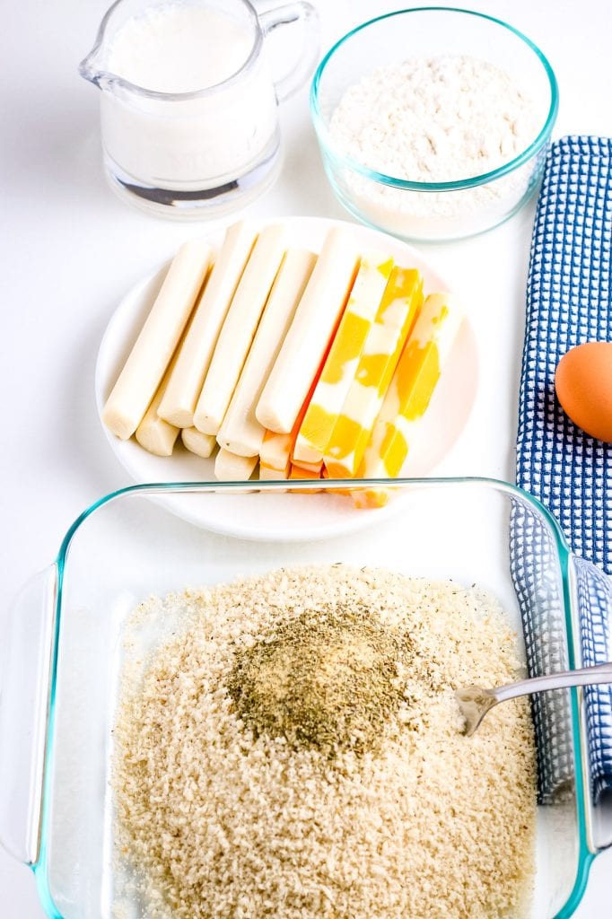 Bread crumbs with seasonings and a spoon in glass baking dish. In background a plate of cheese sticks, dish of flour and glass jar of milk.