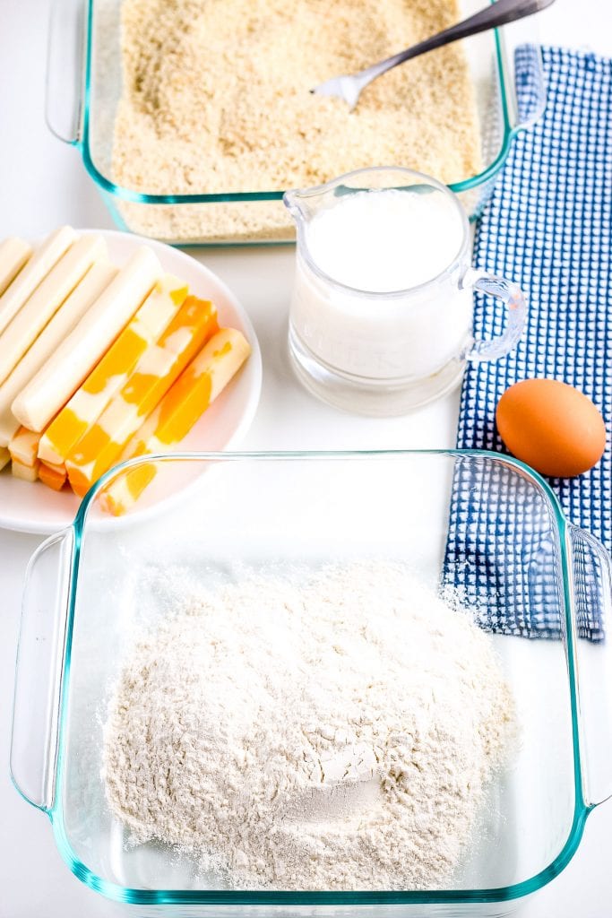 Glass baking dish with flour. In the background a plate of cheese sticks, jar of milk, egg on blue napkin and a glass dish of bread crumbs.