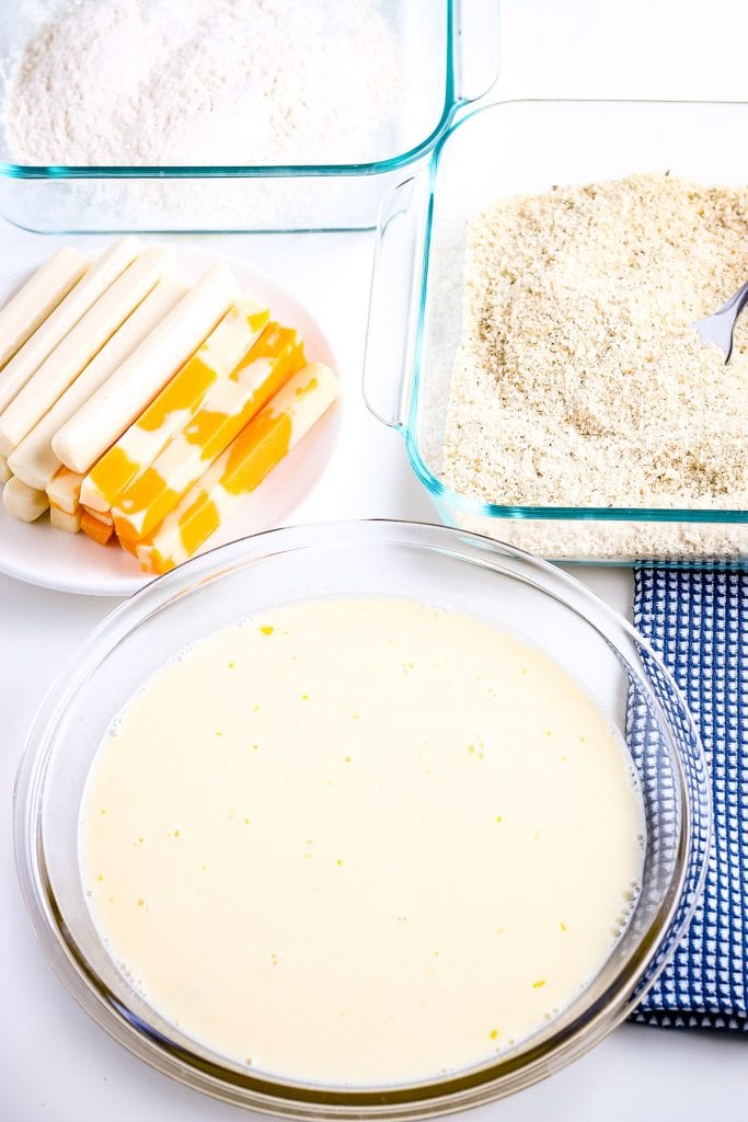 Whisked eggs and milk in glass bowl with cheese sticks, glass container of bread crumbs and flour behind it.