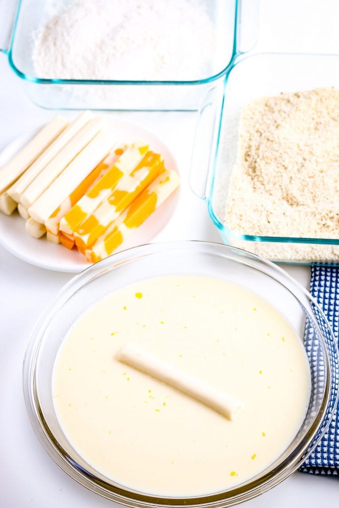 Cheese stick being dipped in egg mixture with bread crumbs in a dish and cheese sticks and flour in a bowl in background.