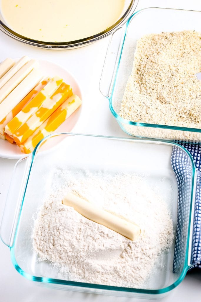 Cheese stick being rolled in flour mixture with cheese sticks, bread crumbs in glass dish and a bowl of mixed eggs in background.