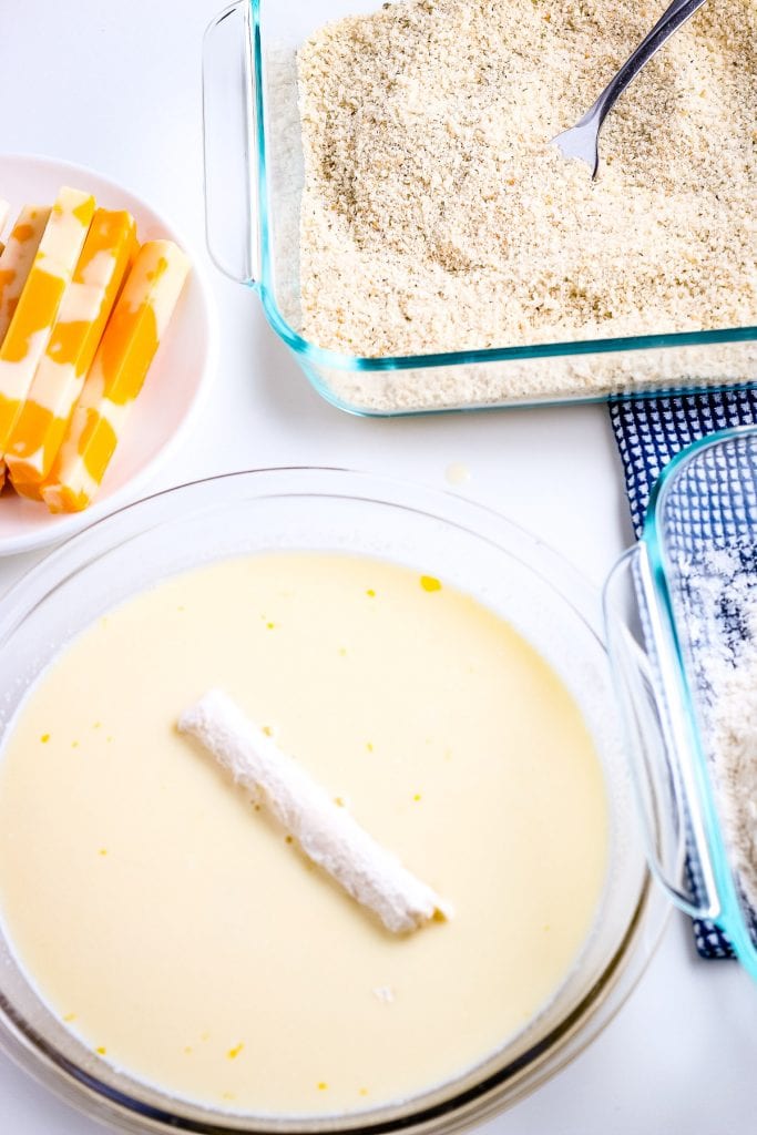 Bread crumb coated cheese stick being dipped in egg mixture with bread crumbs in a dish and cheese sticks in background.