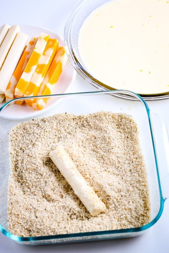 Cheese stick being rolled in bread crumbs in a glass dish with cheese sticks and a bowl of milk in background.