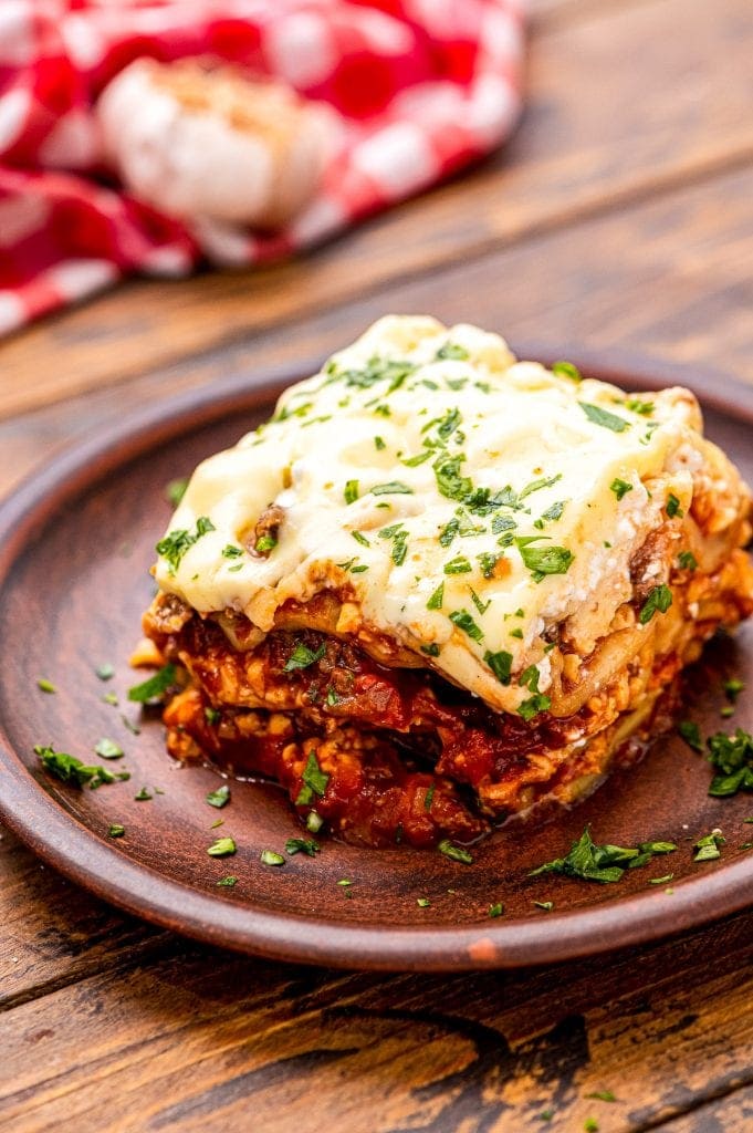 Small wooden plate with a piece of lasagna on it garnished with parsley on wooden background.