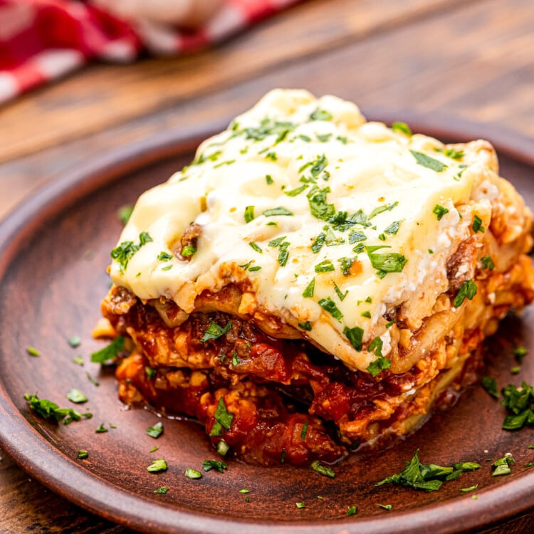 Small wooden plate with a piece of lasagna on it garnished with parsley on wooden background.