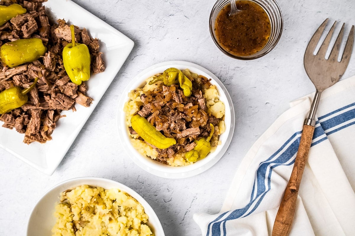 Overhead image of Crock Pot Mississippi Pot Roast in bowl with a platter of it next to it and a bowl of mashed potatoes.