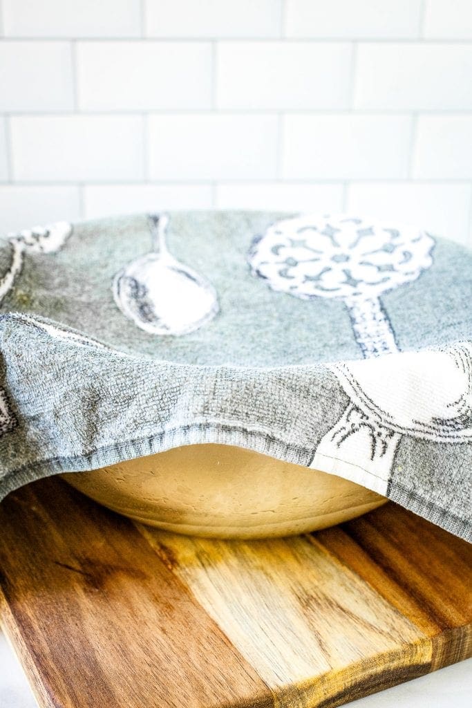 A glass bowl with dough in it covered by a kitchen towel