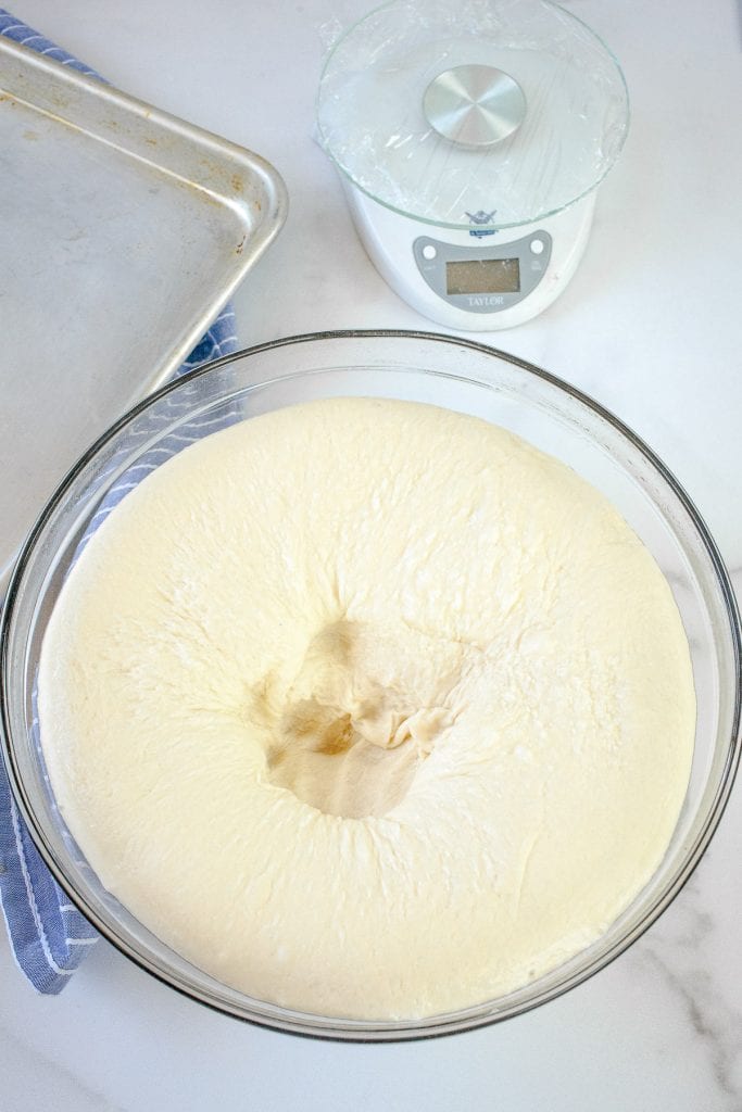 An overhead image of a glass bowl with raised dinner roll dough in it with a fist shape in middle from punching it down.