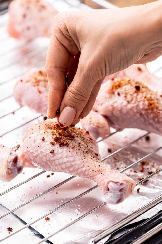 Hand sprinkling seasoning on to raw chicken legs on wire rack sitting on baking sheet.