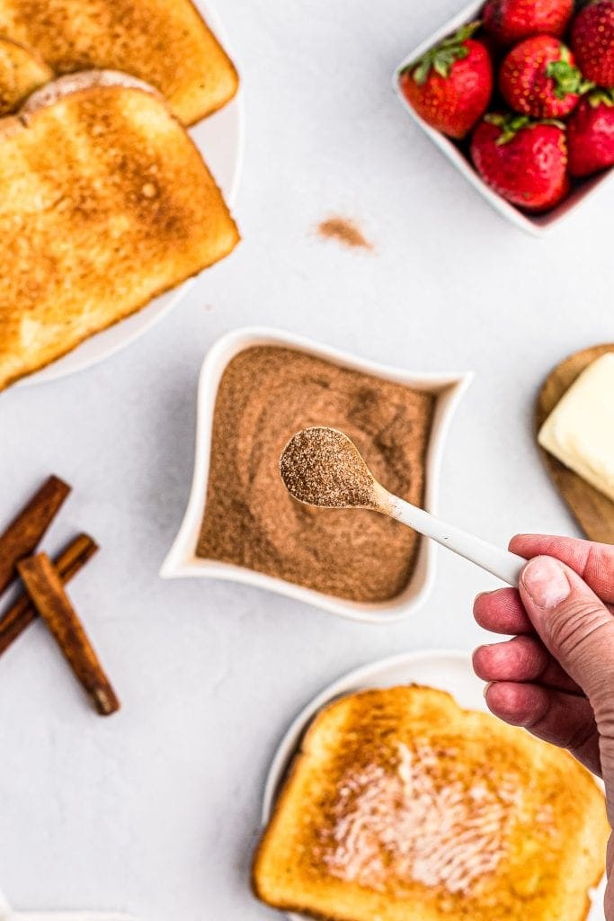 Tiny spoon with cinnamon sugar mixture on it with a bowl of it behind it along with toast and strawberries.