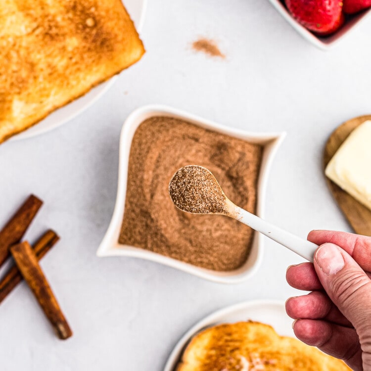 Tiny spoon with cinnamon sugar mixture on it with a bowl of it behind it along with toast and strawberries.