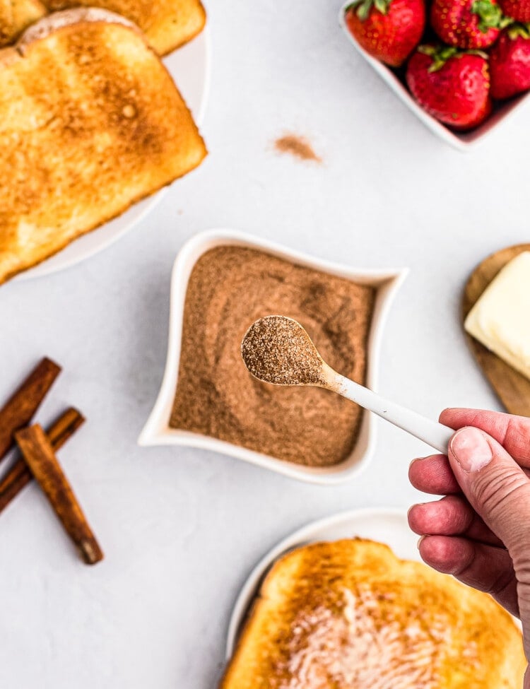 Tiny spoon with cinnamon sugar mixture on it with a bowl of it behind it along with toast and strawberries.