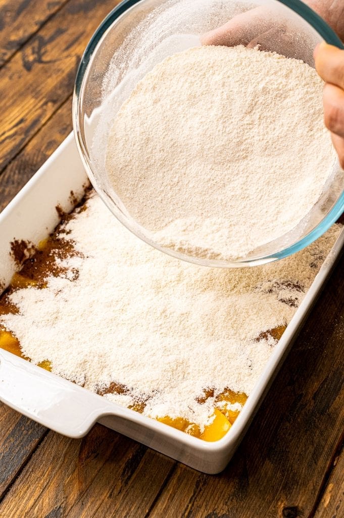 Glass bowl sprinkling cake mix on top of peaches and cinnamon to make dump cake.