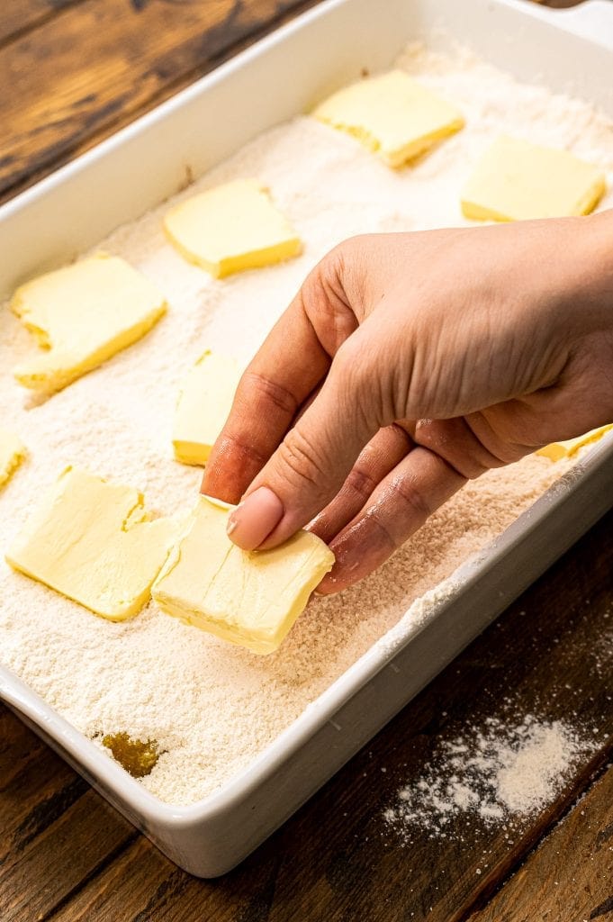 Hand placing slabs of butter on top of cake mix to make dump cake in baking dish.
