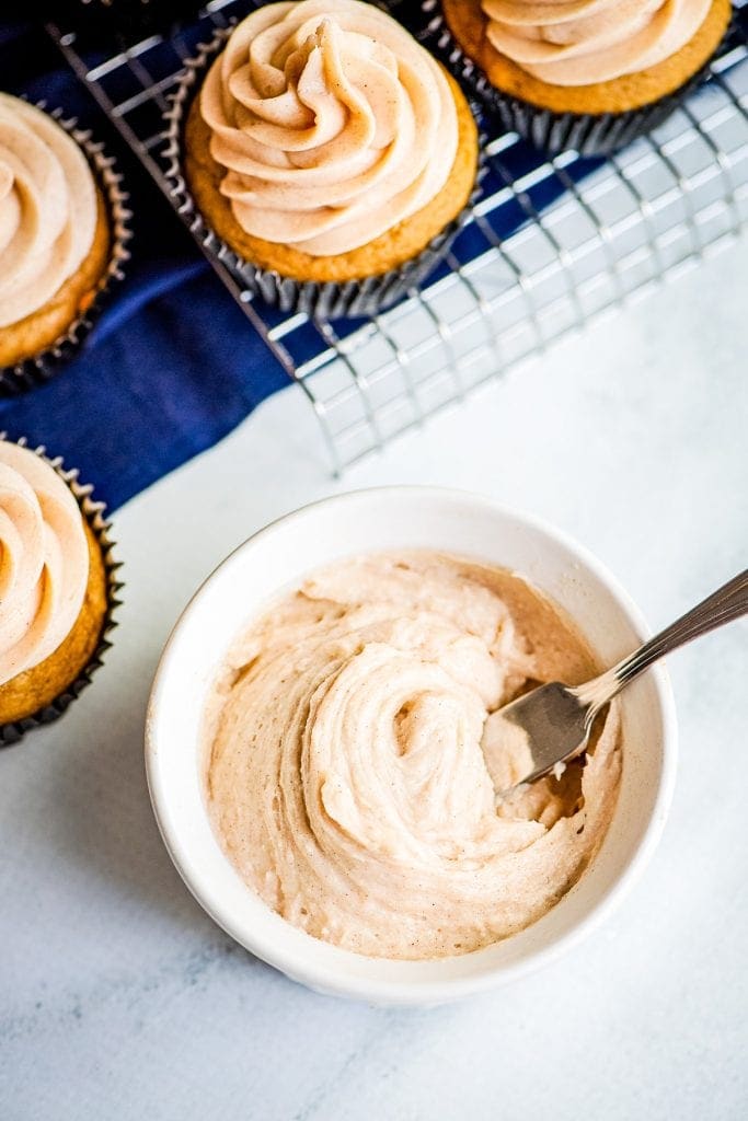 Cinnamon Cream Cheese Frosting in white bowl with spoon in it. Cupcakes in the background that are frosted.