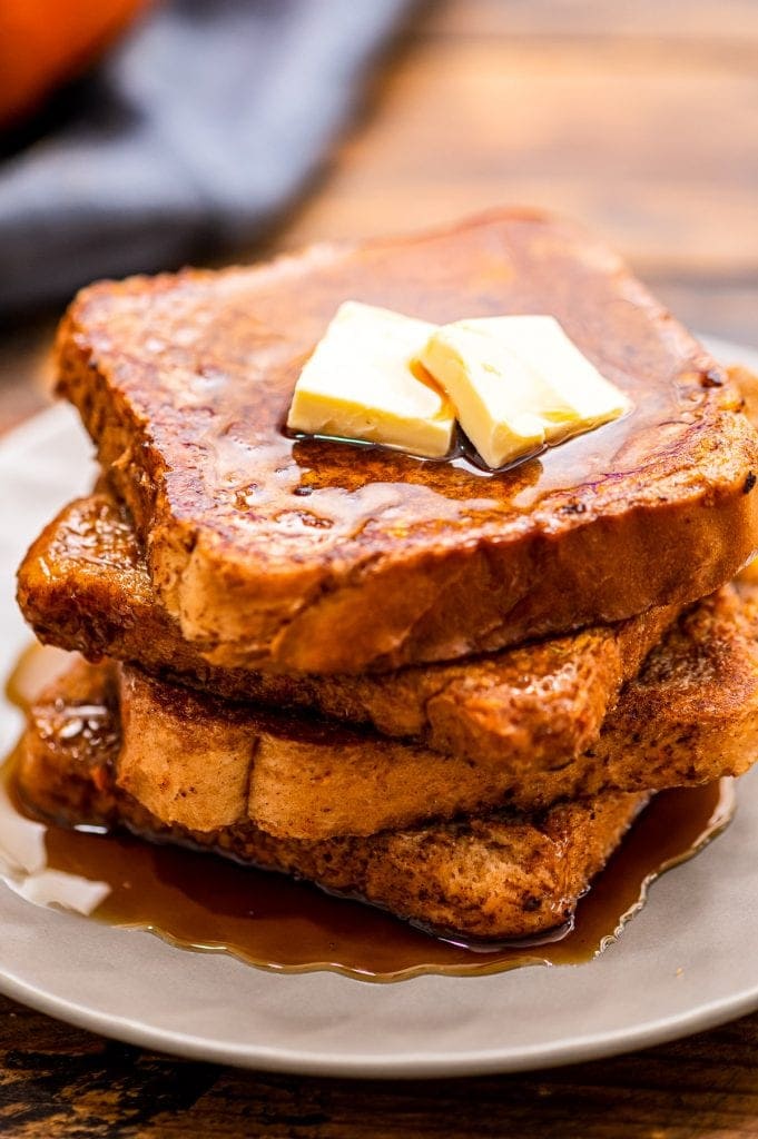 A white plate with a stack of pumpkin french toast topped with butter and syrup