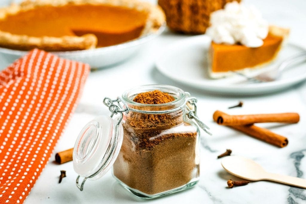 A jar of pumpkin pie spice with pumpkin pie in background